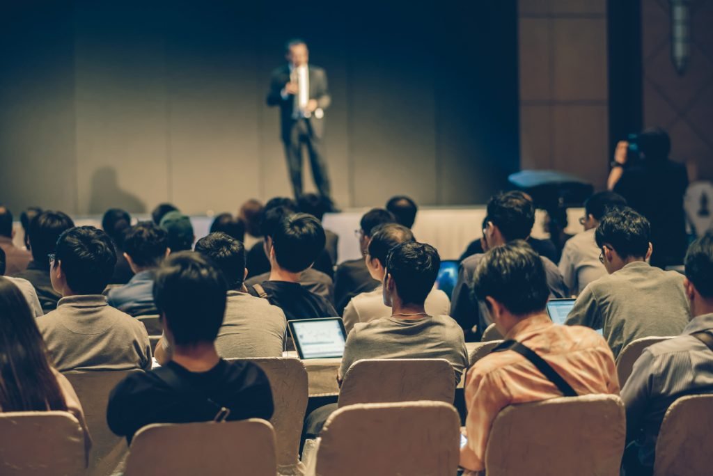 Rear side of Audiences sitting and listening the speackers on the stage in low light conference hall, event and seminar concept